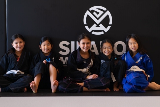 Keira Sammi Emma Allison and Bella sitting up against the Silanoe Martial Arts logo on the wall in the front mat area.