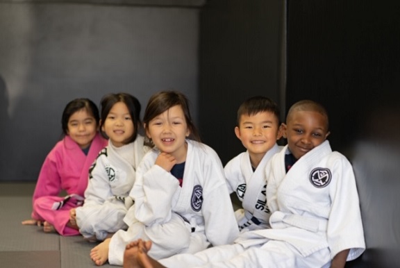 Chelsea, Alayna, Hazel, Colin and Tony from our Kids 1 BJJ program are seated against the wall looking into the camera.