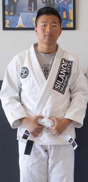 Kalani in a white BJJ gi in front of the picture wall at the San Gabriel Alhambra adjacent Silanoe Martial Arts location.