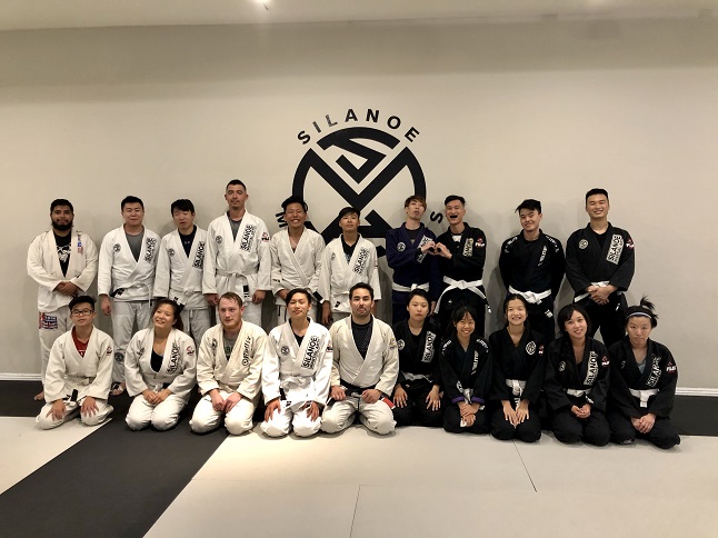 A group picture of students enrolled in the Brazilian Jiu-Jitsu for adults and teens. Standing in the middle on the right hand side in front of the Silanoe logo there is Wah Wu in a black Silanoe Jiu-Jitsu gi uniform.